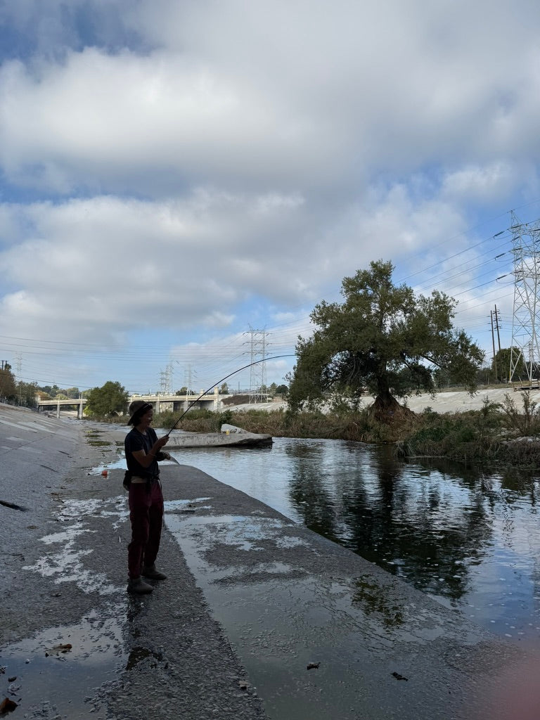 A person is standing on a concrete path next to the LA river as they have a fish hooked on their fly rod, bending their fly rod to a significant degree.